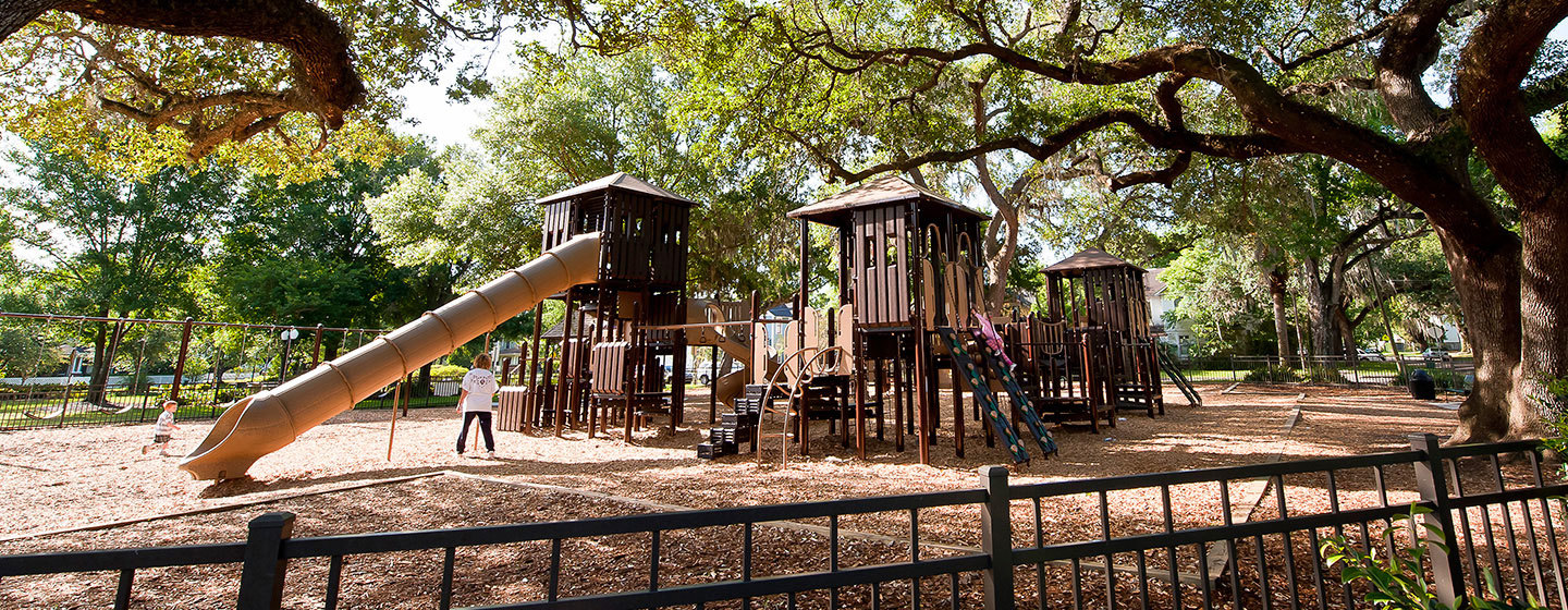 A woodsy image with a brown, wooden-looking playstructure on a woodchip surface with a tan slide on one side and trees all around.
