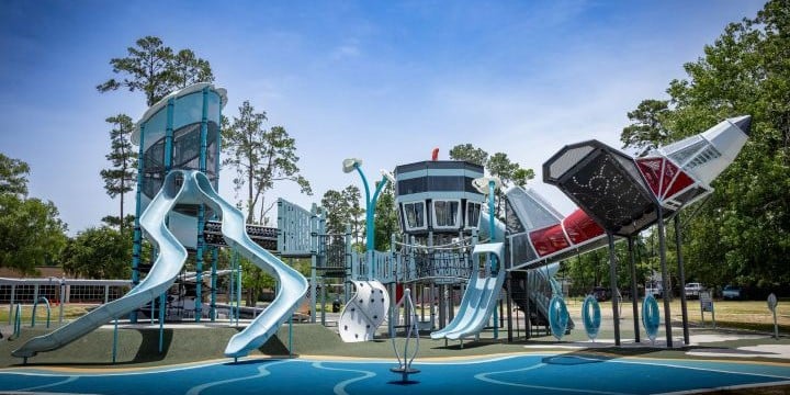 Space themed playground with a red and silver spaceship climber to the right, a space station-themed climber in the middle and a light blue and silver tower to the left with two slides. 
