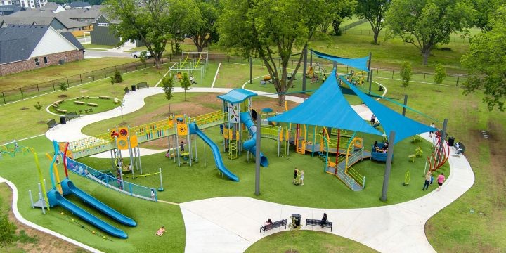 A playground with bright blue shade sails surrounded by a walking path with two blue slides in the foreground and a large climbing structure with several main structures all connected by elevated colorful ramps. 
