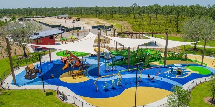 A playground with beige shade sails and bright blue, tan and green surfacing with a green alligator climber and an orange fish climber among other playground components. 