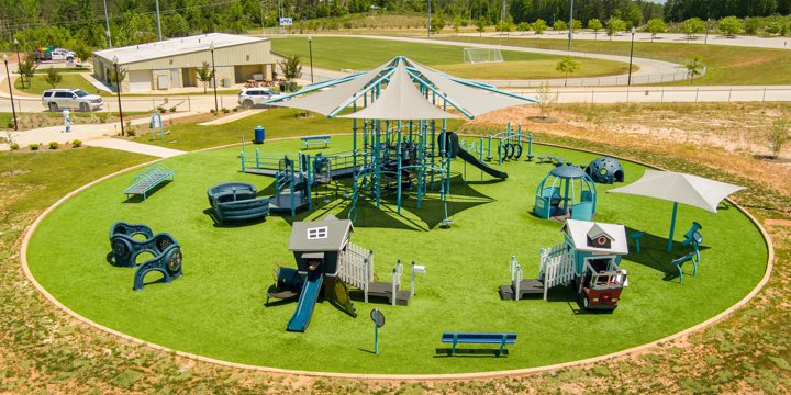 Playground in a circular grassy area with two playhouses in the foreground, multiple freestanding play components and a central rope-net climbing structure with shade sails in the center. 