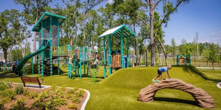A playground with trees in the background and a rock-like arched climber in the foreground. A grassy hill is to the right with two tall blue-green playground structures connected by a bridge on the left. 