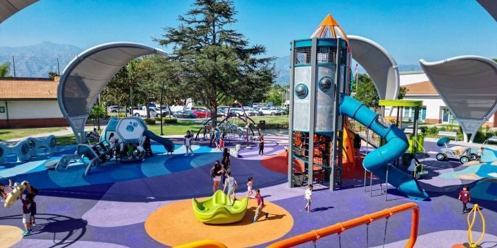 Colorful playground with purple, orange and blue surfacing and a rocket-ship themed custom play structure to the right, surrounded by arching gray shade sails. 