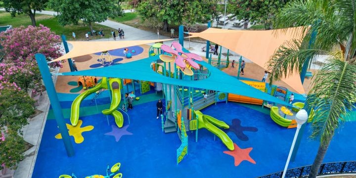 Cartoon-themed playground with blue surfacing and a custom turquoise blue shade structure with flower-shaped roof-toppers poking through. Underneath is a play structure with bright green slides and more. 