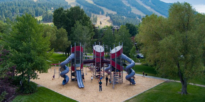 Mountainous scenery with three playground towers in the foreground with internal spiral cable netting, connected by crawl tunnels and two twisty slides. 