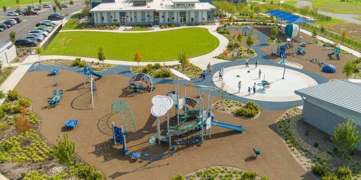 Overhead view of a playground and adjacent splash pad. The playground has a play structure with a cloud-motif roof and multiple inclusive freestanding play structures surrounding it. 