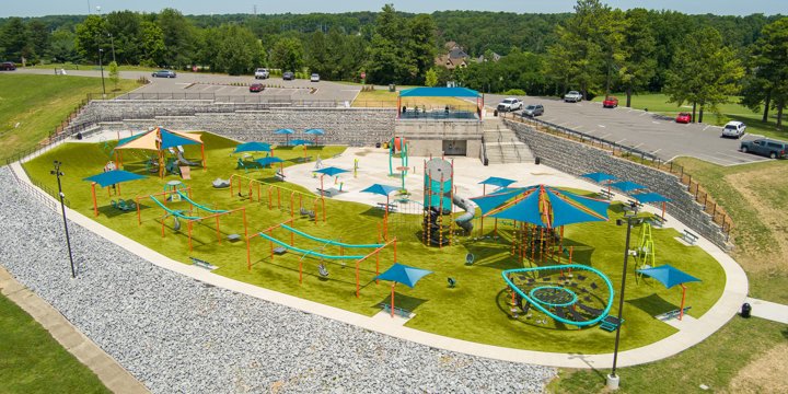 Overhead drone shot of huge oval-shaped play area with "O'"-shaped net climber in the foreground, a tall playground tower in the center, multiple track rides, a net-climbing tower with blue shade. 