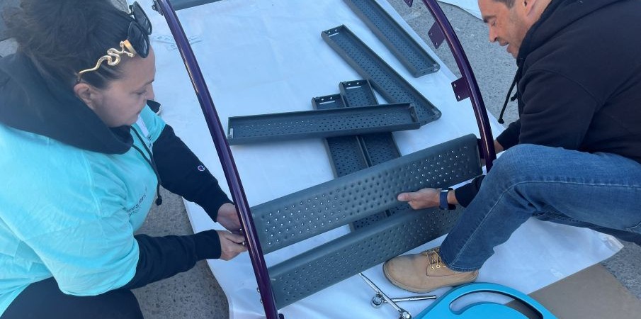 A man and woman crouch on the ground while putting together slats of perforated material to construct a playground component.