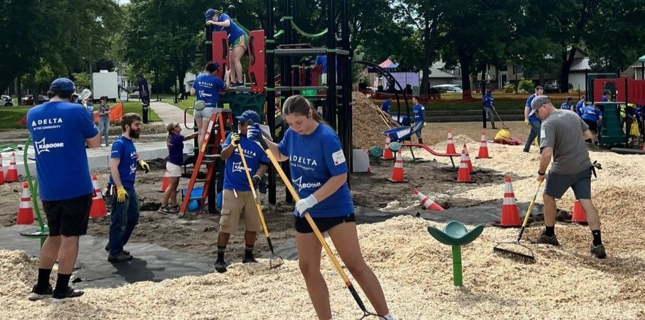 Adults in matching blue t-shirts rake a loose fill playground surface while others consult with each other. A playground is being constructed in the background.