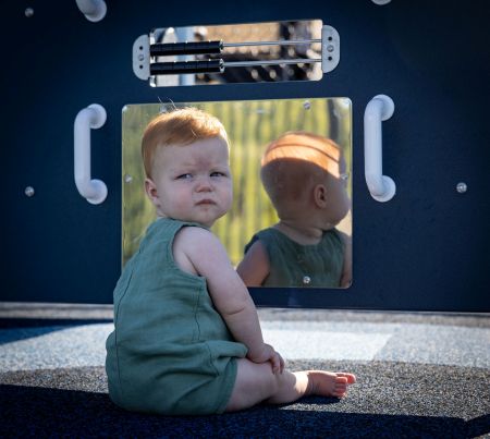 A toddler sits in front of a mirror panel on a playground and looks back at the camera