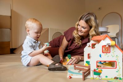 woman lying on her side looking at a toddler holding a toy, with a play house nearby