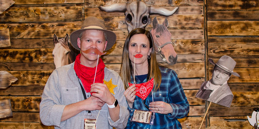 A man and women hold up a fake mustache and lips to their face while standing in front distressed barn wood at a holiday party.