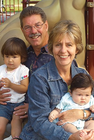 Landscape Structure founders Steve and Barb King smile while sitting with babies on a playground. 