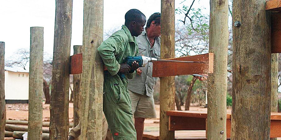 Man drilling into wood plank building a playground while Landscape Structure founder Steve King stands by.