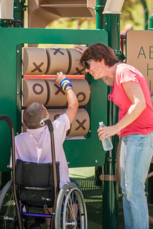 A boy in a wheelchair reaches up to a tic-tac-toe panel, a wheelchair accessible playground equipment component. 