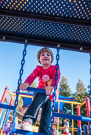 A young girl looks down into the camera as she climbs up a blue playground ladder. 