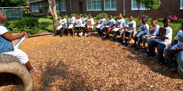 A row of kids sitting on benches face their teacher in an outdoor classroom. 