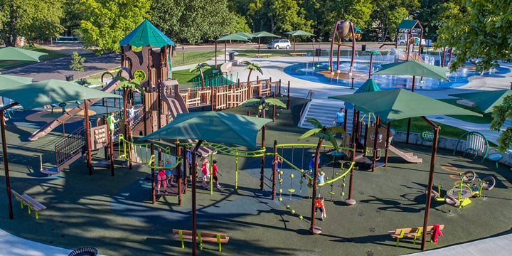 Green SkyWays shade structures and palm tree-shaped posts hover over a brown and green playground with a main brown tower structure in the shape of an old mill with a splash pad in the background. 