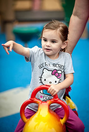 A young girl sitting on a playground rocker points at something. 