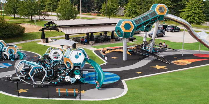 A tall geometric playground tower with a silver slide and other geometric "hedra" playground components in the periphery in the colors of orange and turquoise. 